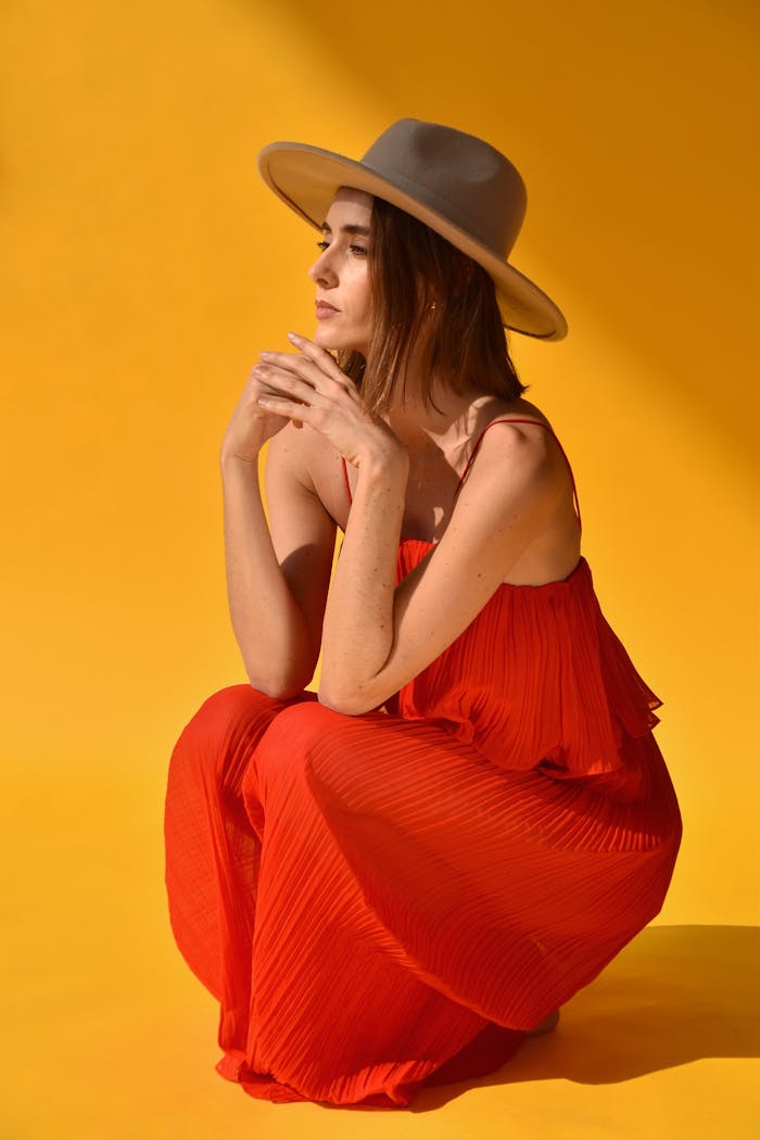 Fashion portrait of a woman in a red dress and hat posing against a vibrant yellow backdrop.